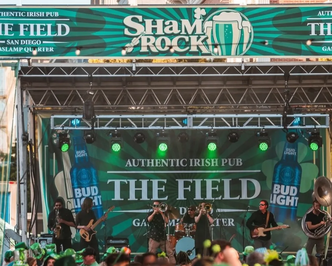 A band performs on an outdoor stage at the ShamROCK festival, with "The Field" and "Authentic Irish Pub" banners and a crowd of attendees in green hats.