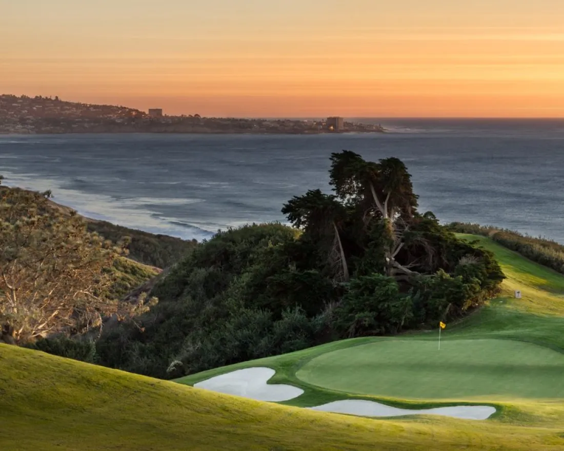 Torrey Pines North Golf Course 15 green with sand bunkers overlooks the ocean at sunset, with distant hills and a coastal town visible in the background.