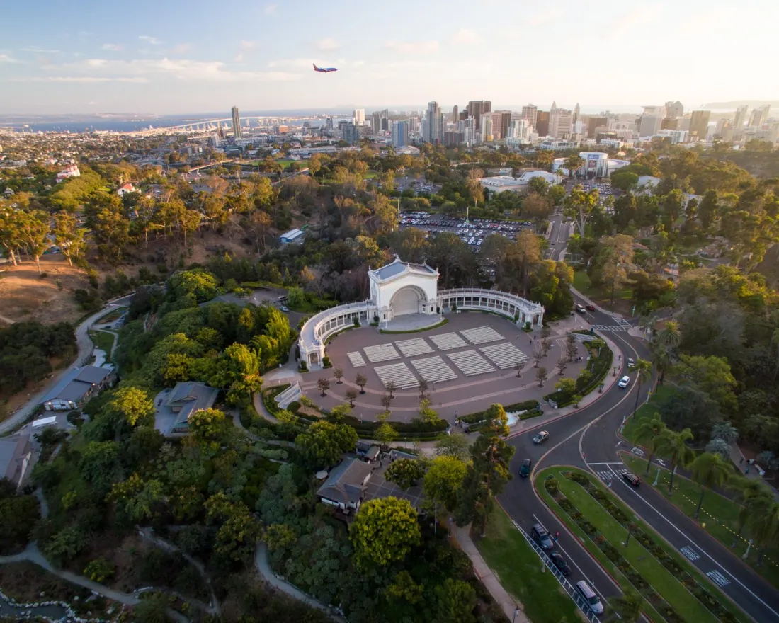 Aerial view of Balboa Park’s Spreckels Organ Pavilion in San Diego, with city skyline and a plane flying overhead in the background.
