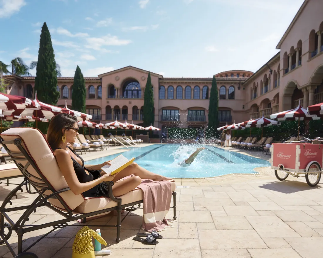 A woman reads a book on a lounge chair by a hotel pool in Del Mar, while someone dives into the water and red-and-white umbrellas line the poolside.