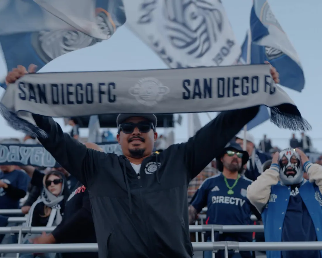 A San Diego FC soccer support holds a team banner aloft as fans dance and cheer at beautiful Snapdragon Stadium in San Diego.
