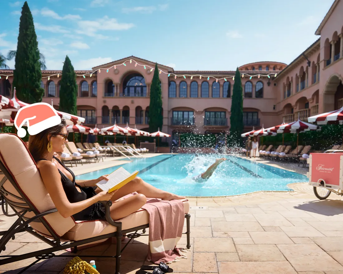 A woman wearing a Santa hat reads by a hotel pool while someone dives in, with red and white umbrellas and Mediterranean-style buildings in the background.