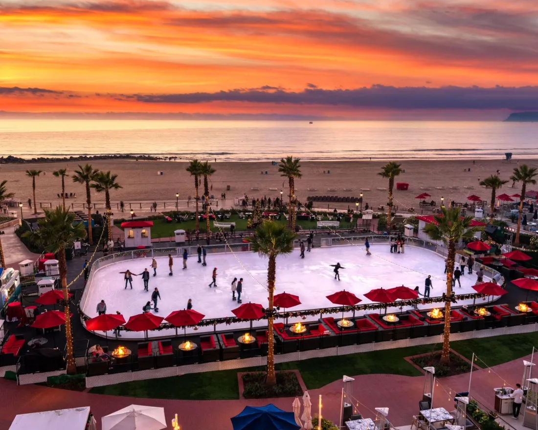 An outdoor ice skating rink by the beach at sunset, with people skating, palm trees, red umbrellas, and the ocean in the background.