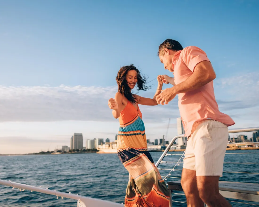 Two people dance together on the deck of a boat with the San Diego skyline and blue sky in the background.