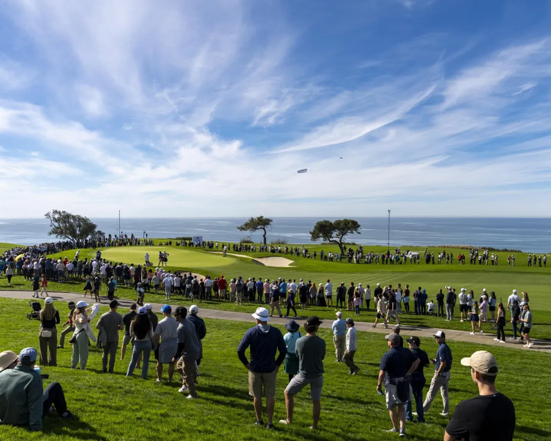 Wide shot of the Famers Insurance open with crowds watching one of the players putt on the green under blue skies and with views of the Pacific Ocean