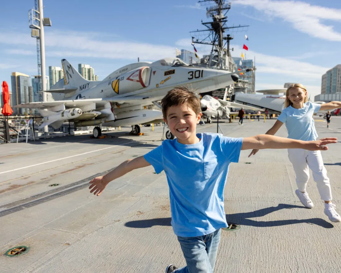 Two children in blue shirts pose with arms outstretched on the deck of the USS Midway Museum aircraft carrier, with a Navy jet and city buildings in the background.