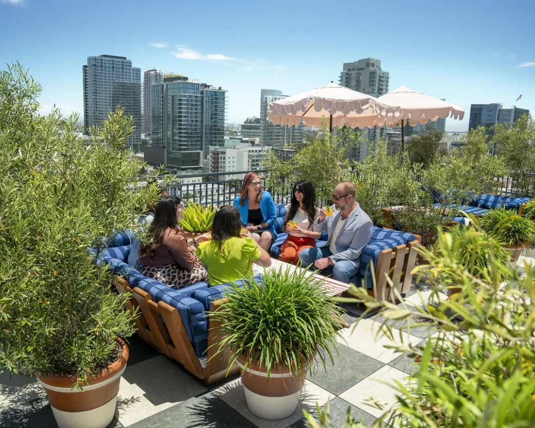 Five people sit and talk on a rooftop patio surrounded by plants, with San Diego city buildings in the background and a pink umbrella providing shade.