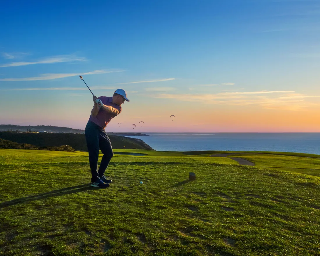 A golfer swings a club on a green golf course overlooking the ocean at sunset, with a clear sky and a few birds in the distance.