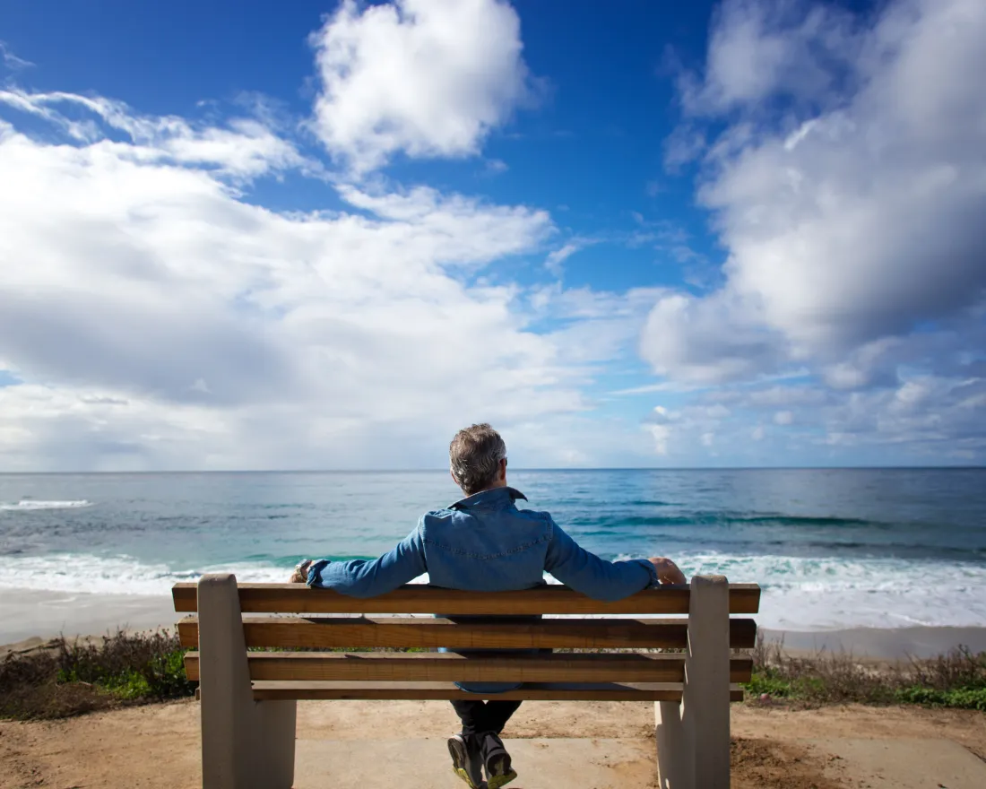 A person sits alone on a bench facing the ocean under a partly cloudy sky.