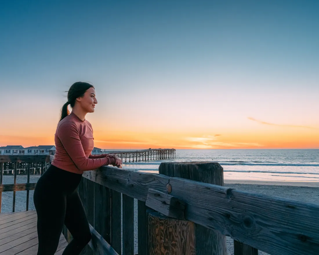 A solitary woman stands on a pier looking at a beach near sunset in San Diego.