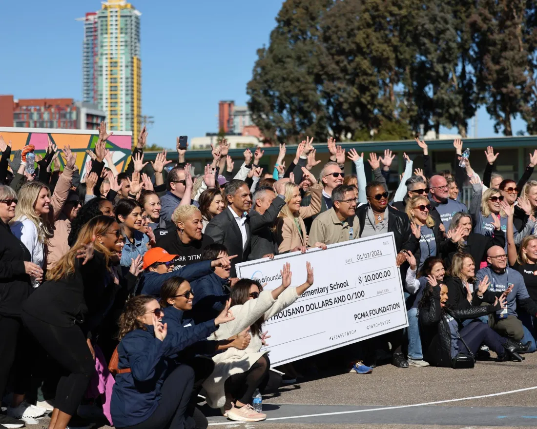 Large group of adults outdoors at a corporate volunteer or charity event, smiling and raising their hands as they pose together around two oversized donation checks held at the center of the group, with city buildings and trees in the background.
