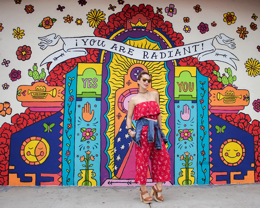 Alt text:   Colorful mural on the exterior wall of Artlexia, a Hispanic-owned business in San Diego, featuring a radiant, shrine-like design with bold, bright colors, flowers, hearts, candles, eyes, and smiling suns. A large banner at the top reads “YOU ARE RADIANT!” with “YES” and “YOU” on either side of a central glowing figure. In front of the mural, a person in a red, off-the-shoulder dress with a denim jacket tied around their waist stands on the sidewalk, looking confidently at the camera.