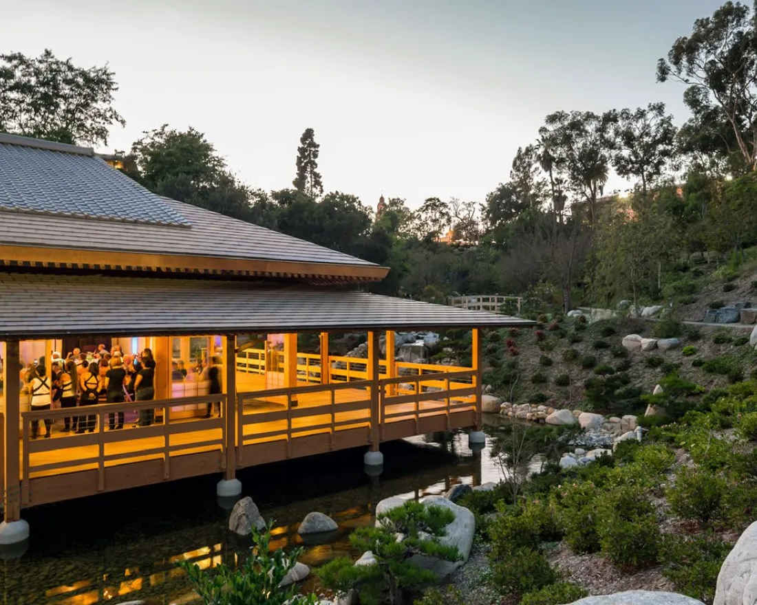 Evening view of an AANHPI cultural gathering inside a warmly lit wooden pavilion at the Japanese Friendship Garden in Balboa Park, San Diego, surrounded by stones, water, and lush hillside greenery.