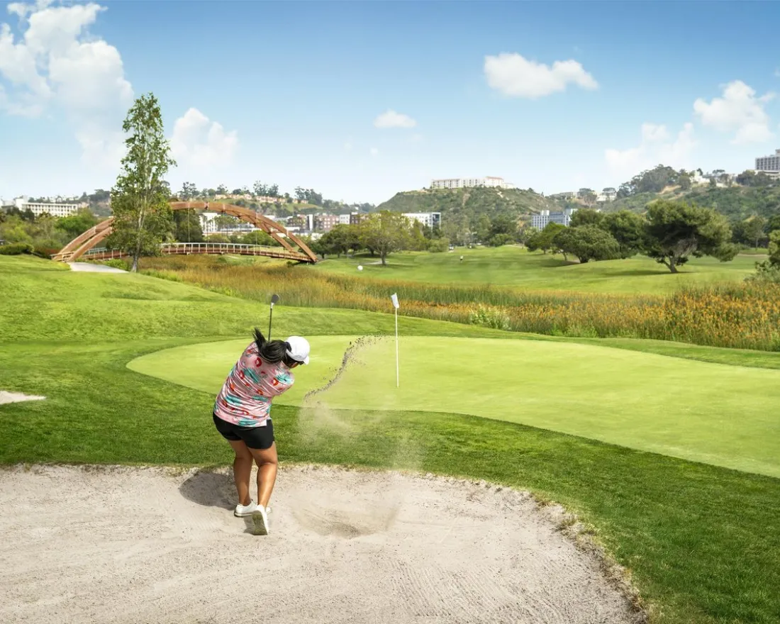 A golfer in shorts and a pink shirt drives a golf ball out of a sand trap toward the green on a sunny day in San Diego's beautiful Mission Valley.