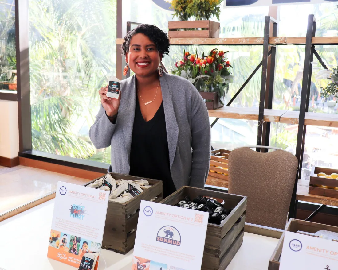 Smiling entrepreneur standing behind a display table for a Black-owned small business in San Diego, holding up a packaged product. The booth features wooden crates filled with neatly arranged bath or skincare items, promotional cards highlighting local Black-owned brands, and large windows in the background showing palm trees and a sunny Southern California landscape.