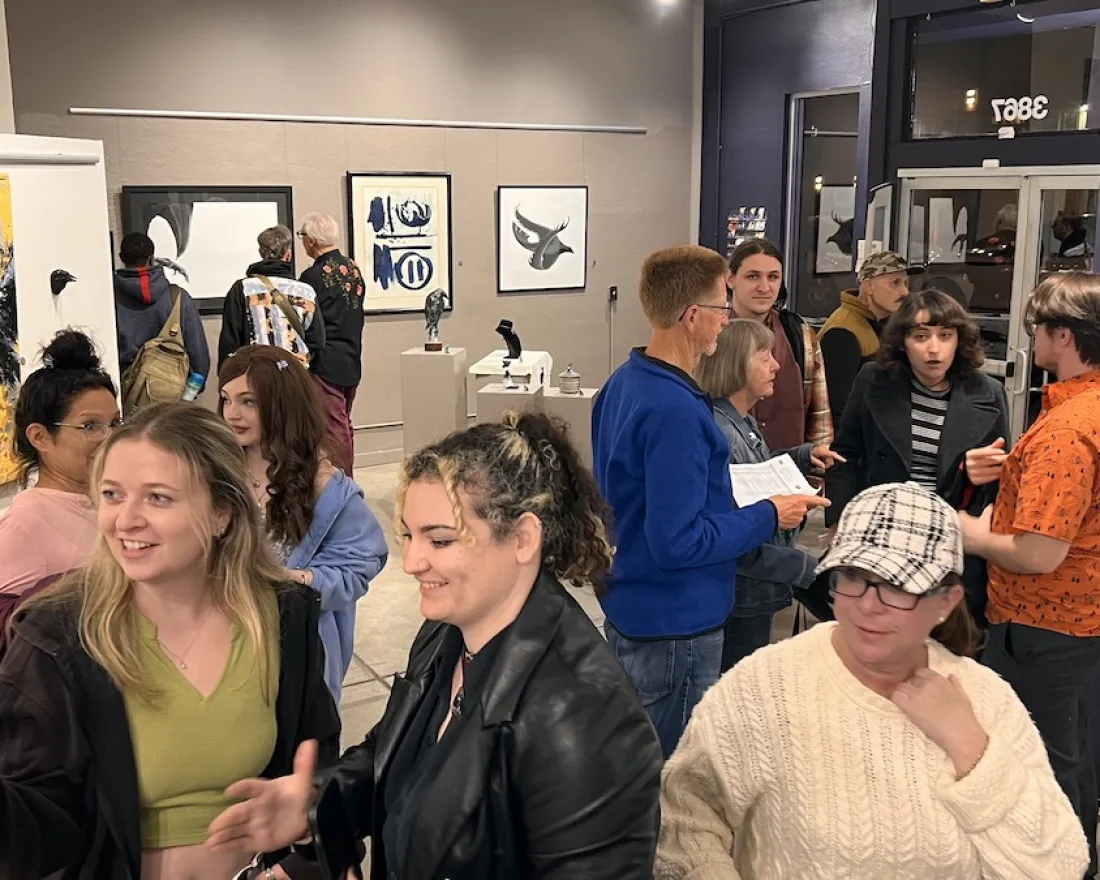 A diverse group of people mingle and chat inside The Studio Door art gallery in Hillcrest, San Diego, surrounded by colorful contemporary paintings and sculptures. Small clusters of guests sit and stand close together, smiling, talking, and looking at artwork on the walls, creating a warm, welcoming atmosphere that reflects LGBTQIA+ community event spaces and gatherings in San Diego.