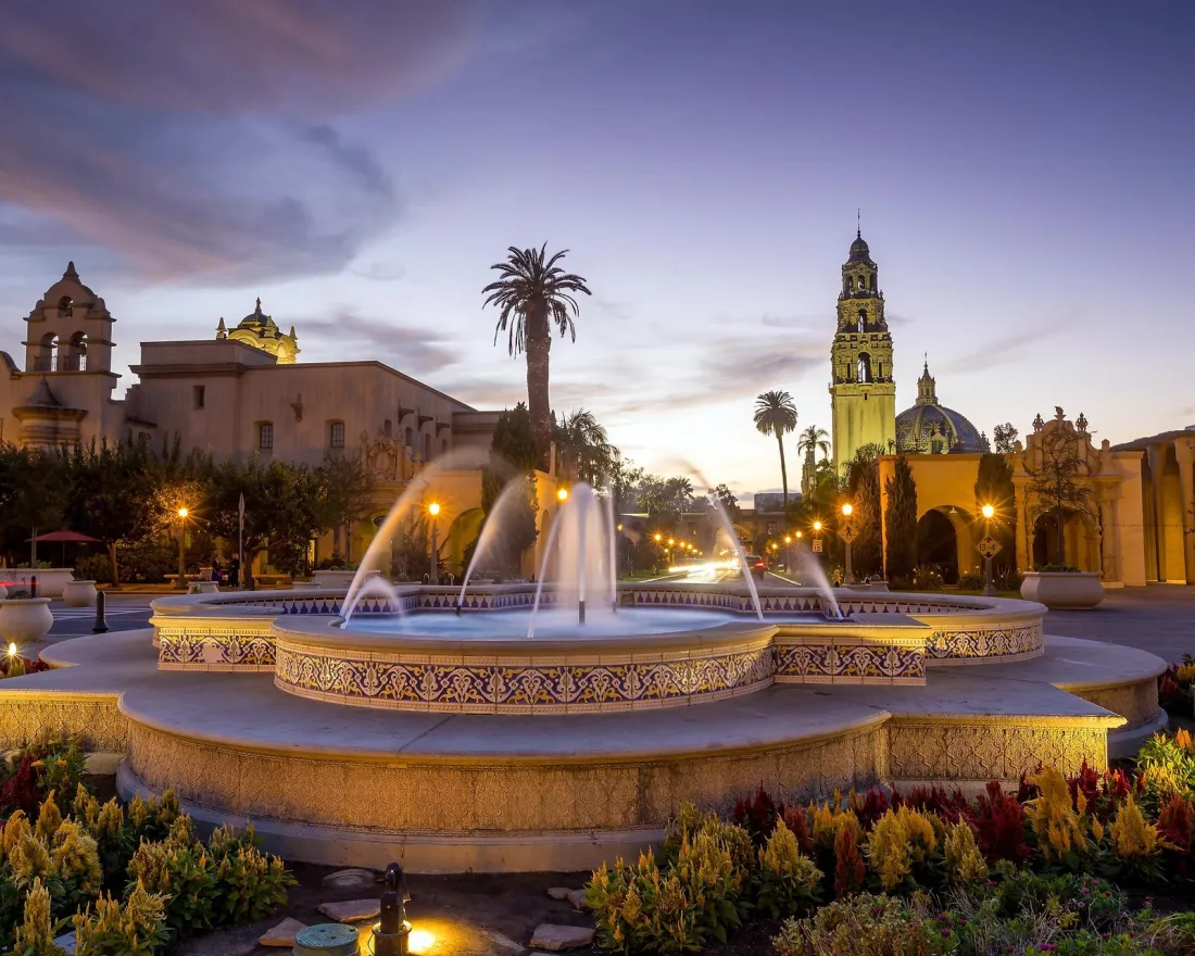 Balboa Park Fountain surrounded by vibrant flowers at dusk, with historic buildings and a clock tower in the background.