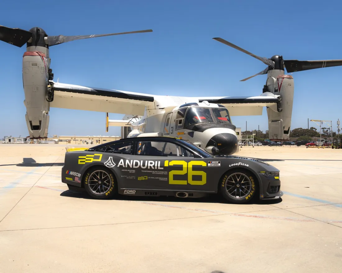 A sleek black NASCAR racing car with the number 26 is parked in front of a U.S. Navy Osprey aircraft on a sunny day at Naval Base Coronado near San Diego.