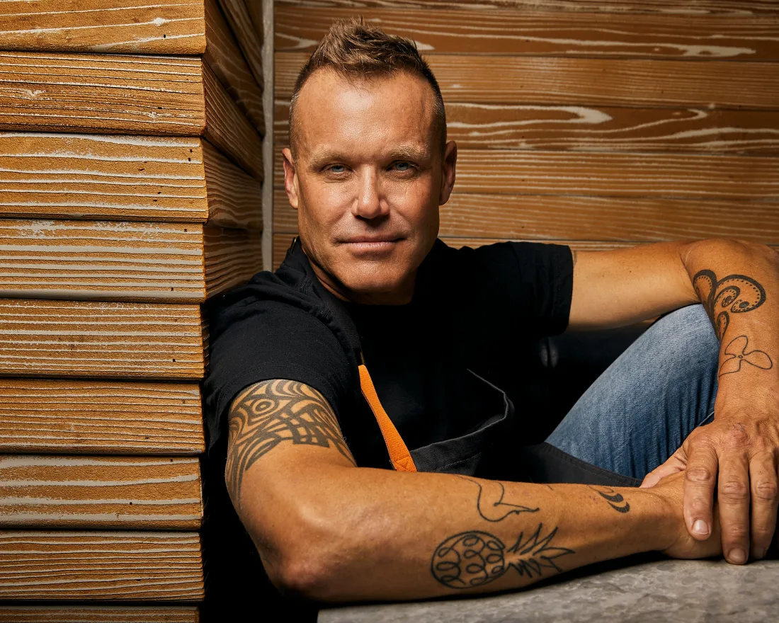 Man with tattoos seated next to wooden panels, looking confidently at the camera.