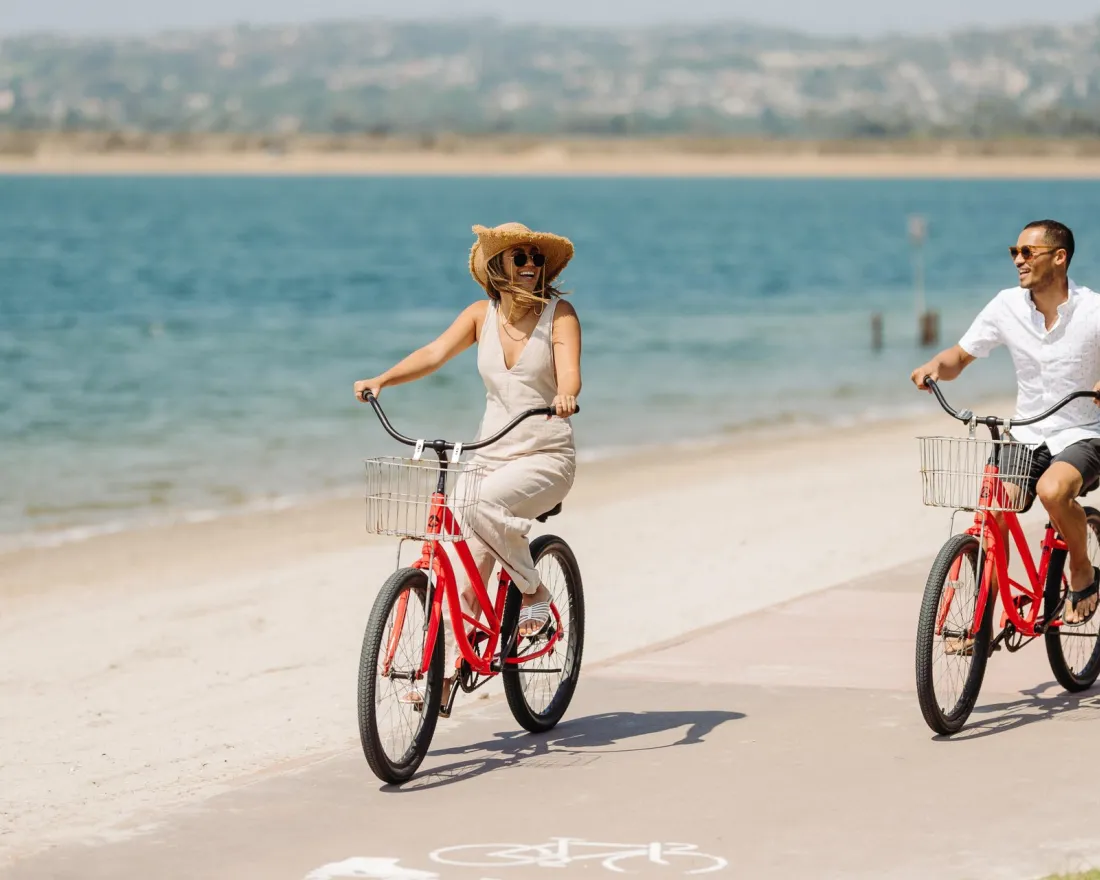 A man and woman ride red bicycles along a Mission Bay's sandy beach by the water.