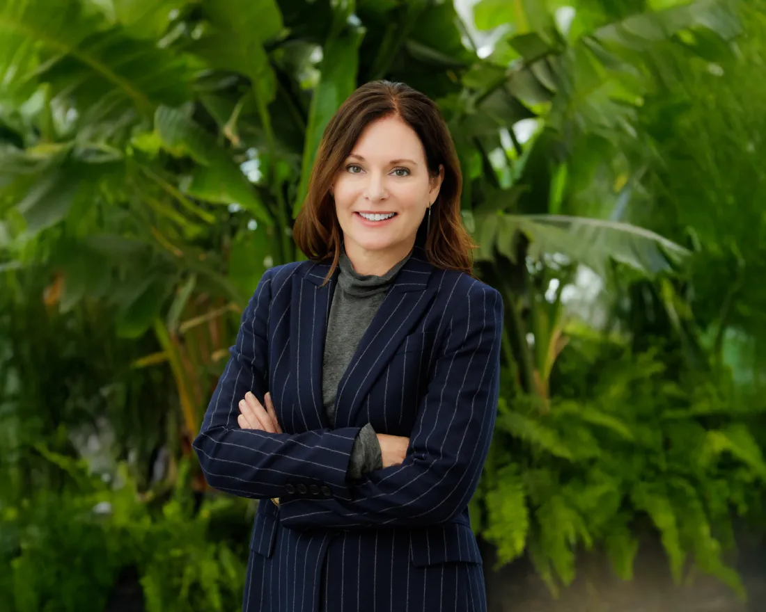Kerri Kapich in a striped blazer stands confidently with arms crossed among lush green plants.