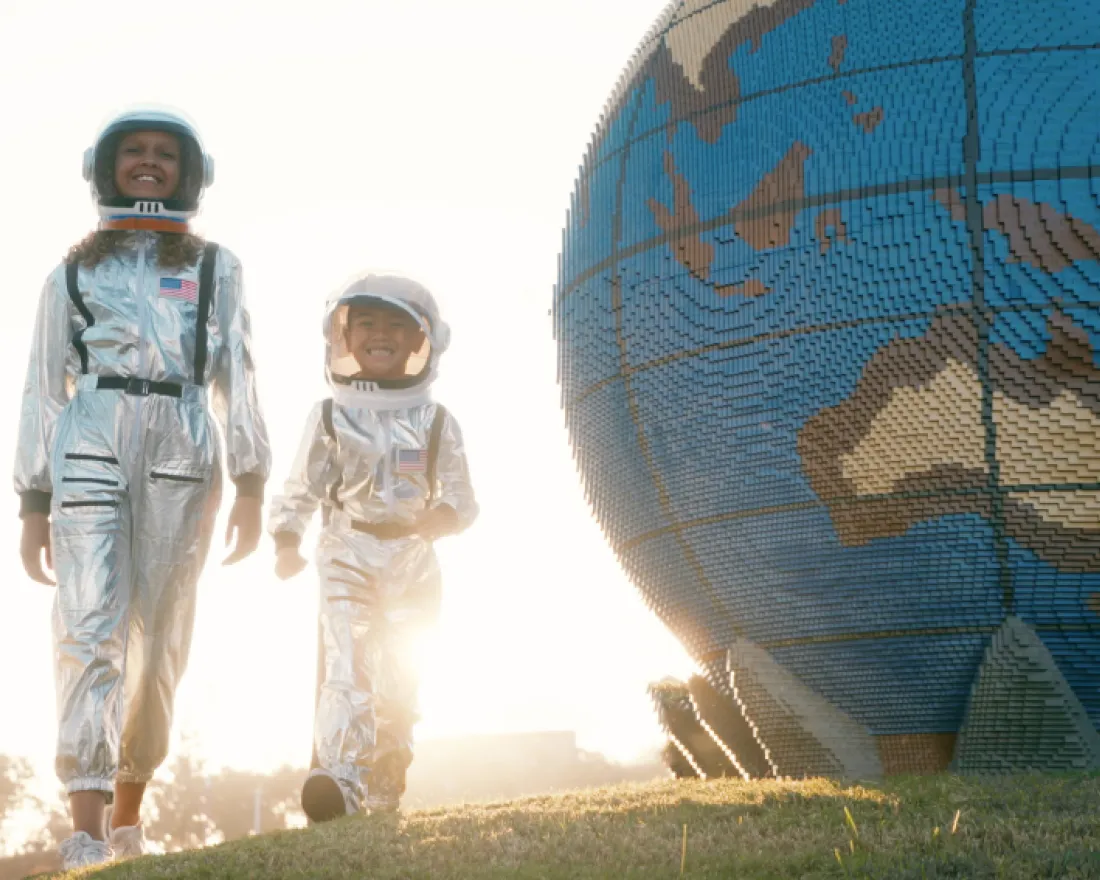 Two child astronauts walk beside a large globe sculpture at sunset.