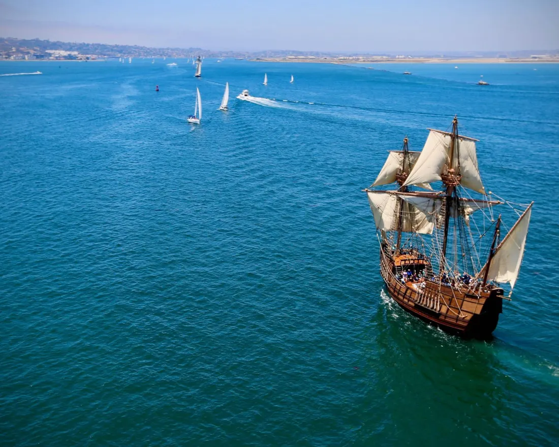 San Salvador wooden sailing ship replica with white sails navigates through a turquoise sea, with smaller boats nearby.