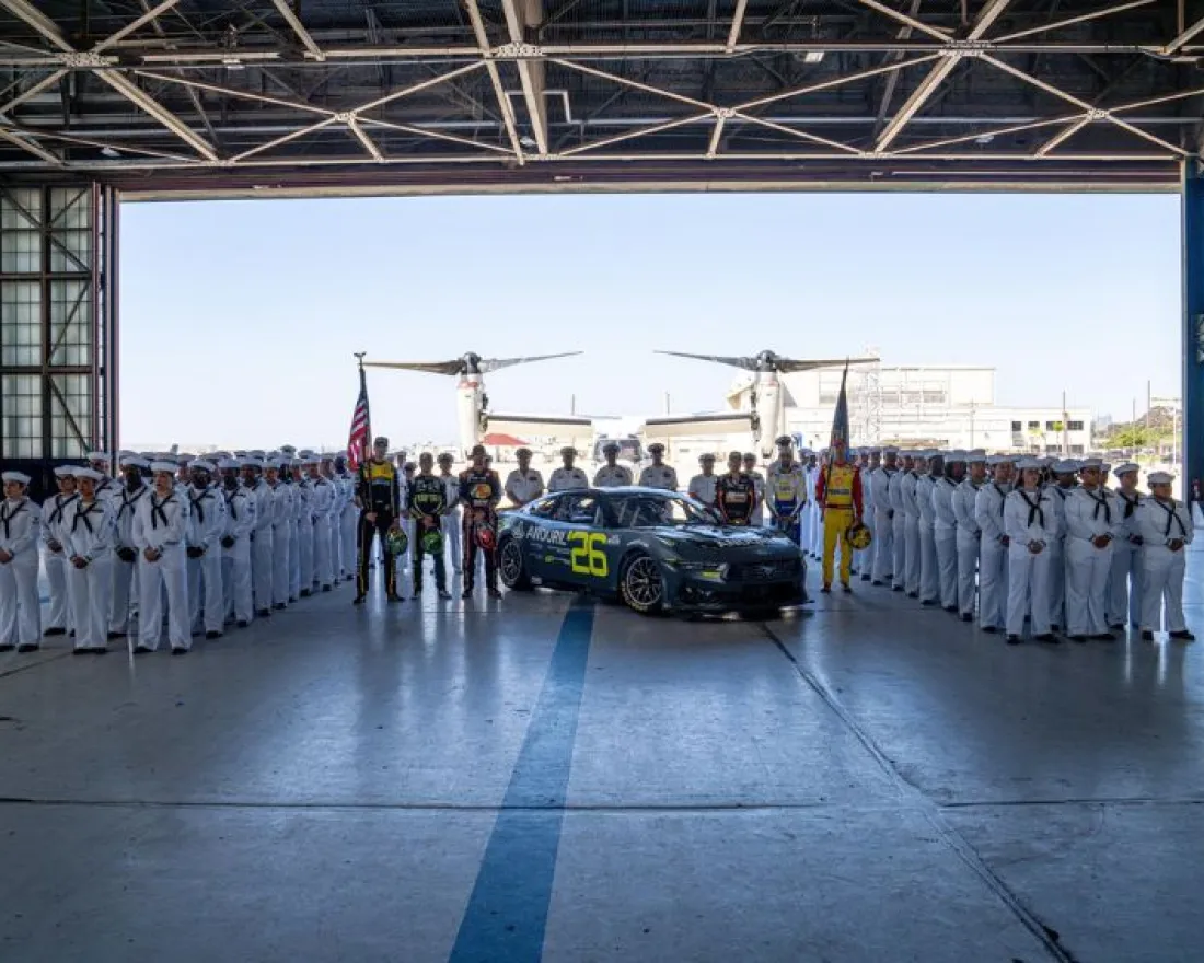 Naval personnel and aircraft gather inside a hangar, with a car prominently featured in the center.