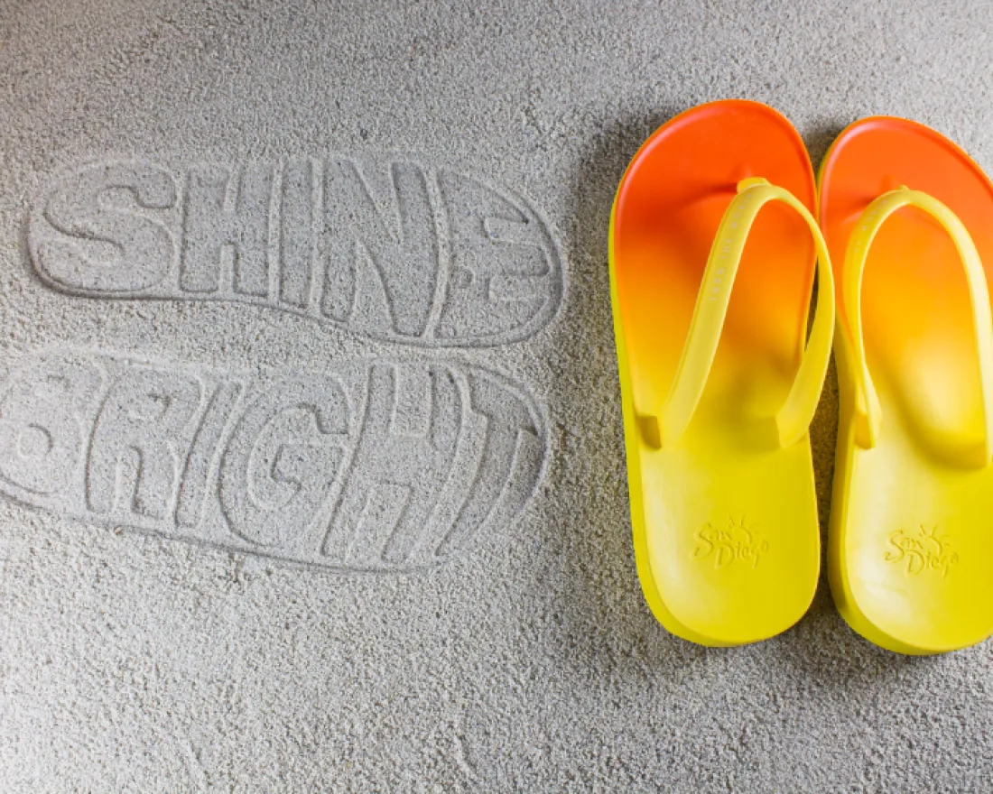 Yellow and orange slippers beside the words "SHINE BRIGHT" imprinted in sand.