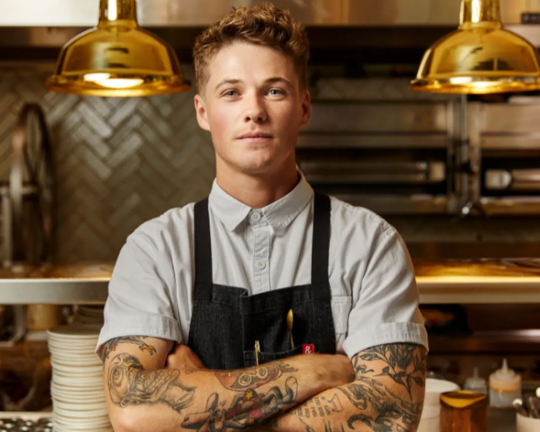 A tattooed chef stands confidently in a modern kitchen, arms crossed under warm lighting.
