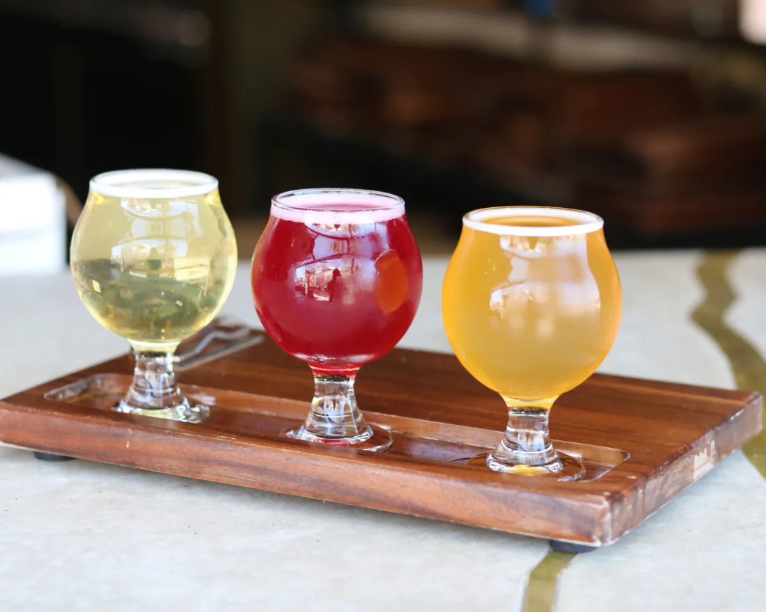 Three small glasses of different colored cider from Biouvac Ciderworks in San Diego on a wooden tasting paddle.
