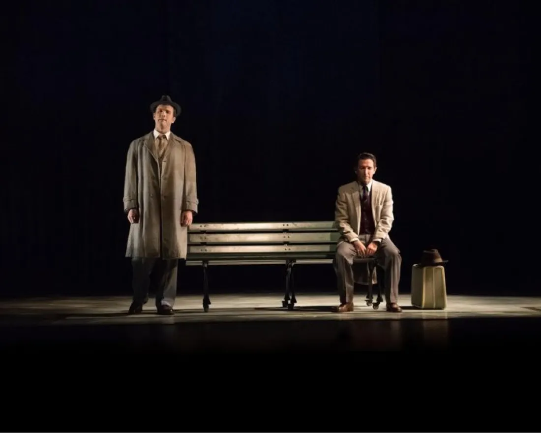 Two actors on stage, one standing in a trench coat and the other sitting on a bench.