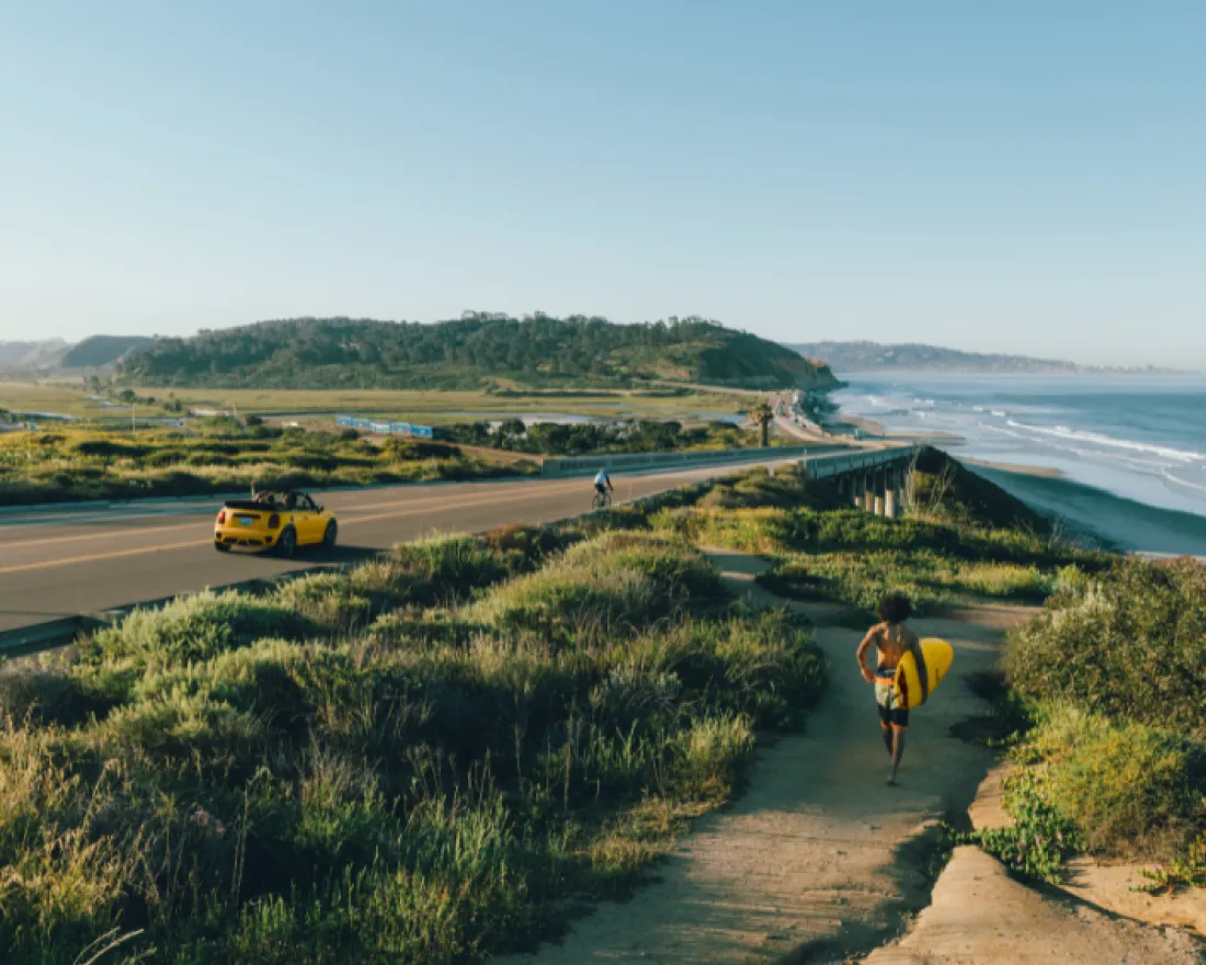 A person walks along a path by the shore, with waves and a coastline view.