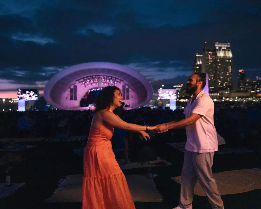 A couple dances joyfully at an outdoor concert at the Rady Shell, with the nighttime San Diego skyline illuminated in the background.