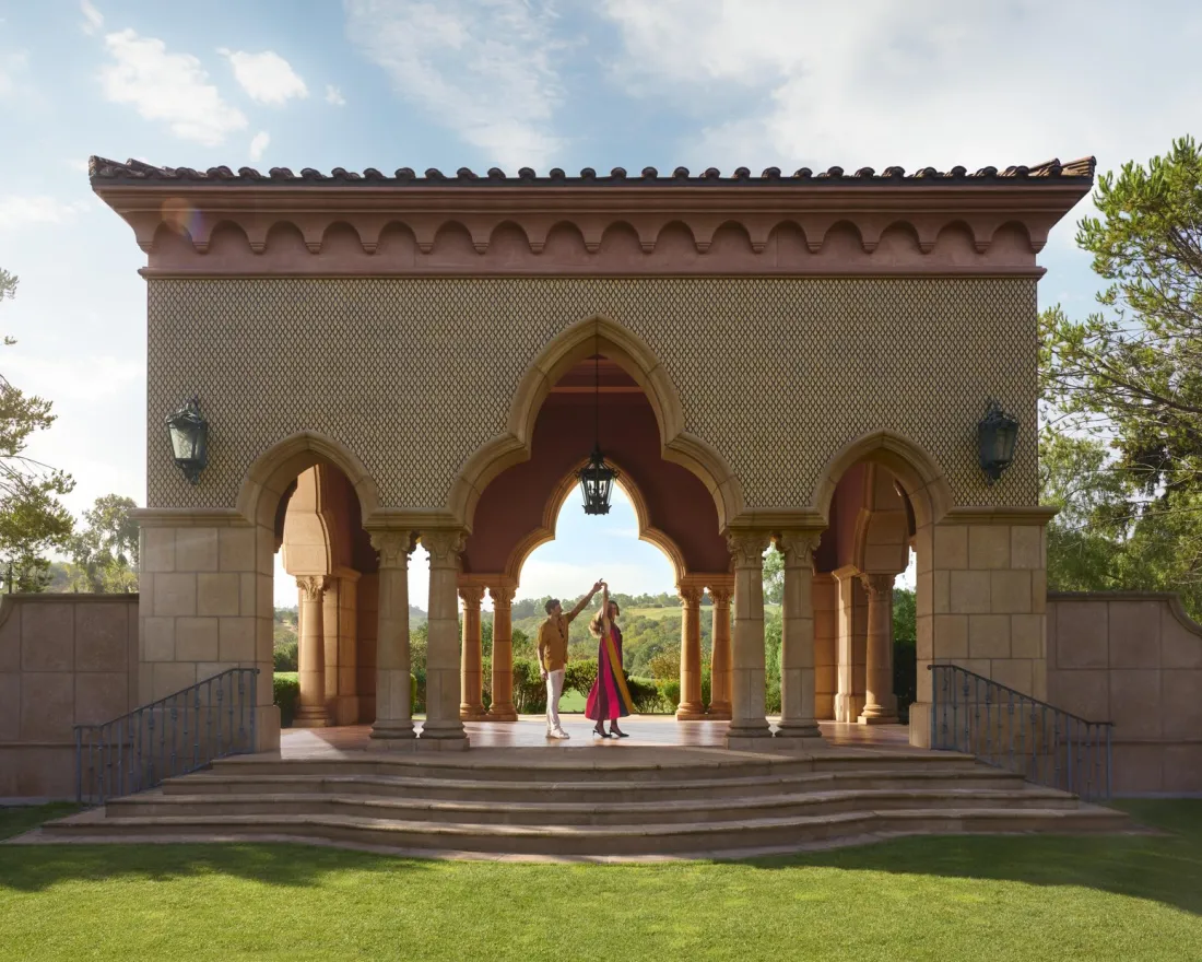 A couple stands on the steps of a Moroccan-style pavilion at the Fairmont Grand Hotel in San Diego, surrounded by greenery.