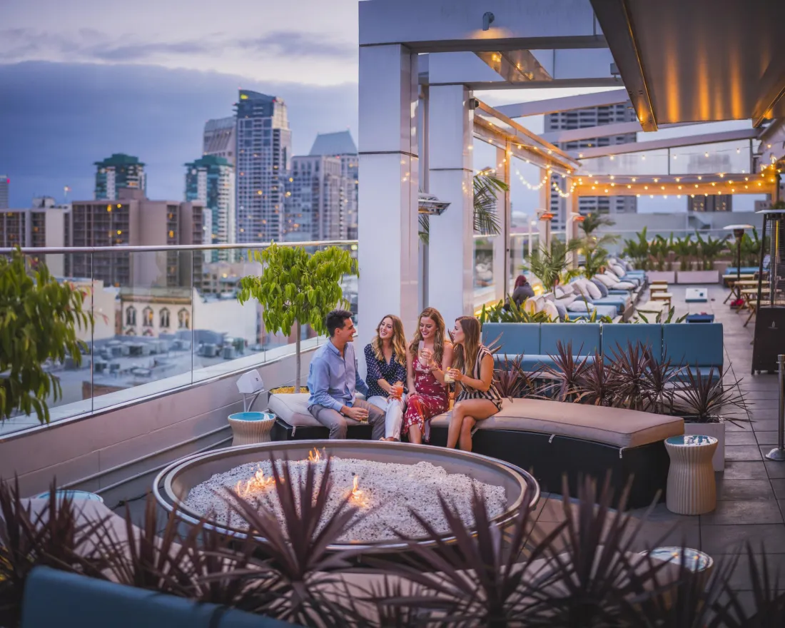 Group of friends chatting around a fire pit on a rooftop with the San Diego skyline in the background.