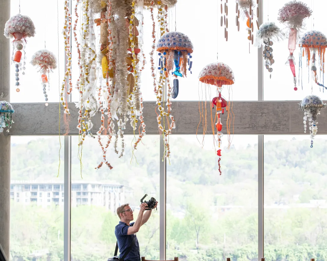 A person photographing hanging jellyfish sculptures in a brightly lit room at the Washed Ashore exhibition at the San Diego Natural History Museum in Balboa Park.
