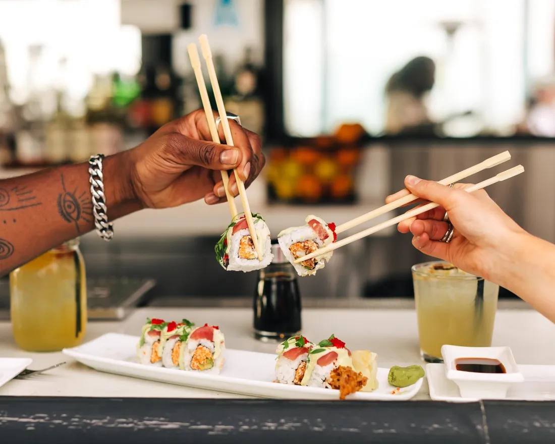Two hands using chopsticks to share sushi rolls at a restaurant table in San Diego.