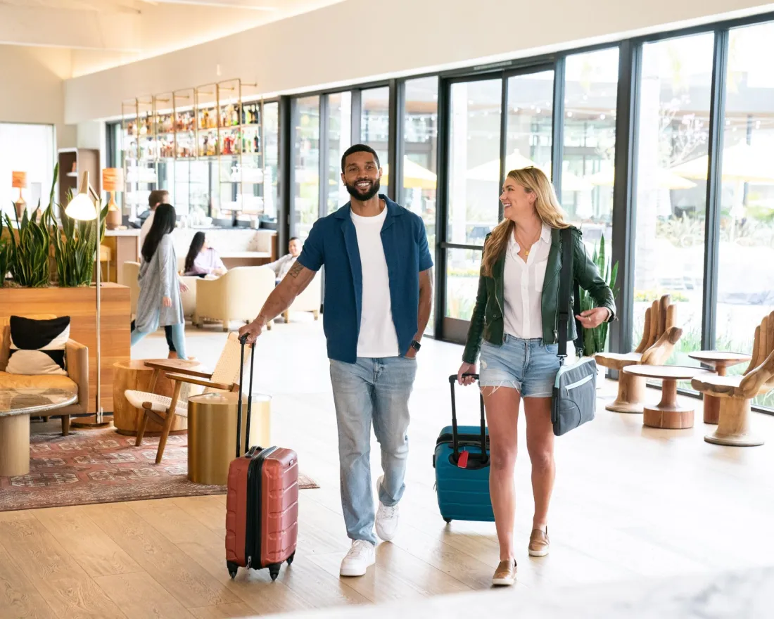A man and woman walk through a modern hotel lobby at the Town and Country Resort in San Diego, each pulling luggage.
