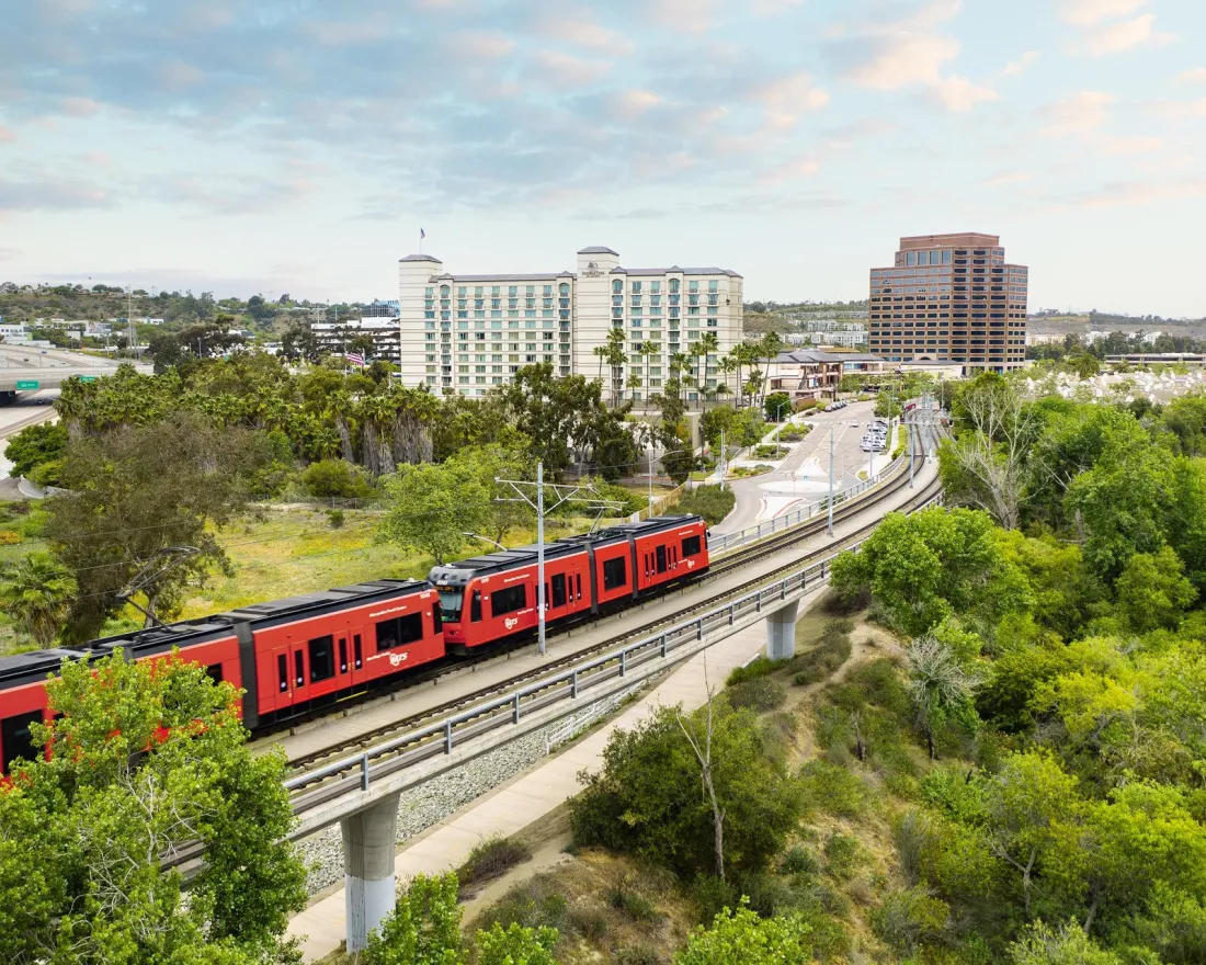 A red San Diego Trolley train travels along tracks near buildings and greenery under a cloudy sky in the UTC/La Jolla area of San Diego.