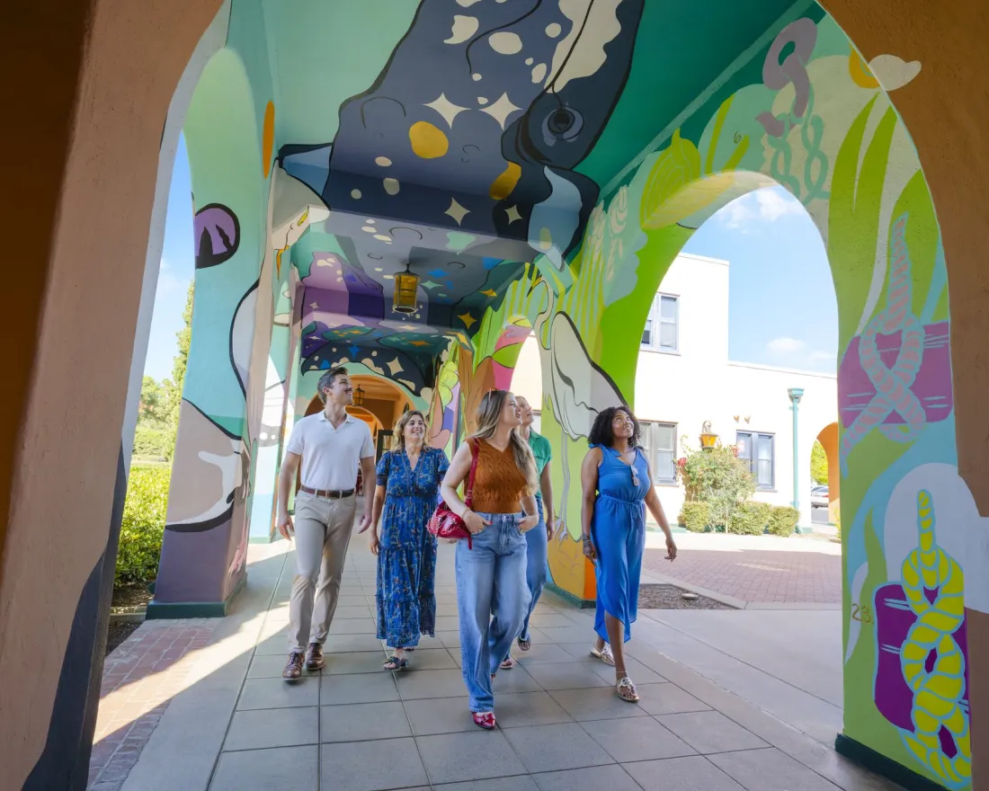 A diverse group of five people walks through a colorful, mural-covered archway at Arts District Liberty Station in San Diego.
