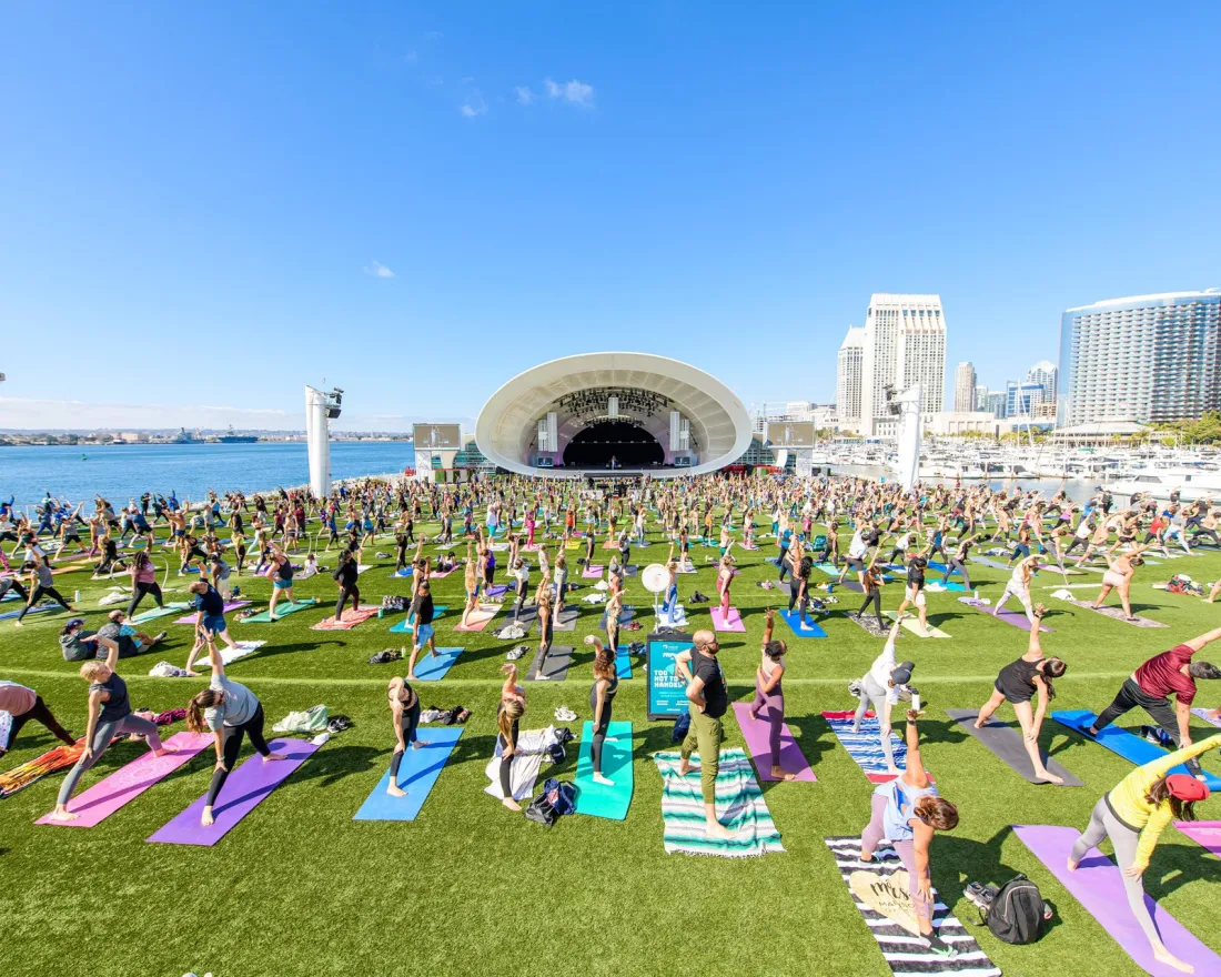 A large group of people practicing yoga on colorful mats outdoors near a water body at They Rady Shell at Jacobs Park.