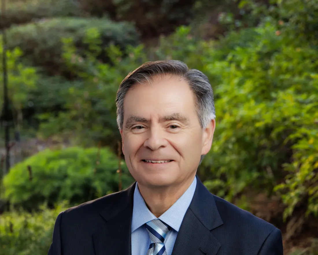 Rick Meza, CFO, in a suit stands outdoors, smiling, with greenery in the background.
