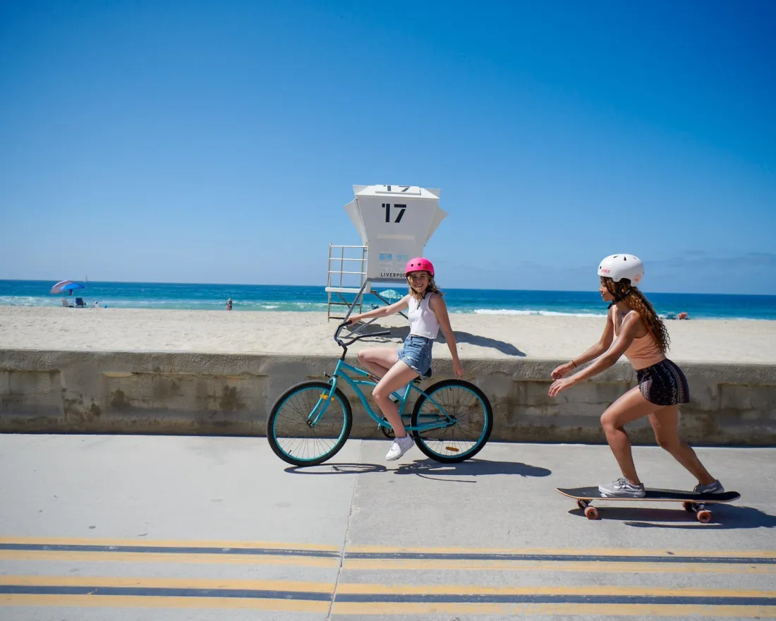 Two girls enjoying a sunny beach day along the Mission Beach Boardwalk in San Diego: one on a bicycle and the other on a skateboard.