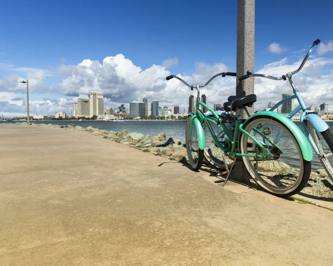 Two teal bicycles leaning against a pole by the San Diego Bay waterfront with the city skyline in the background.