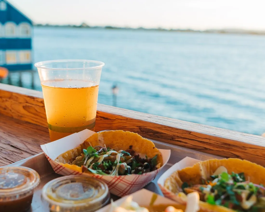 Two taco bowls with toppings, sauces, and a beer, set against a waterfront view in San Diego.