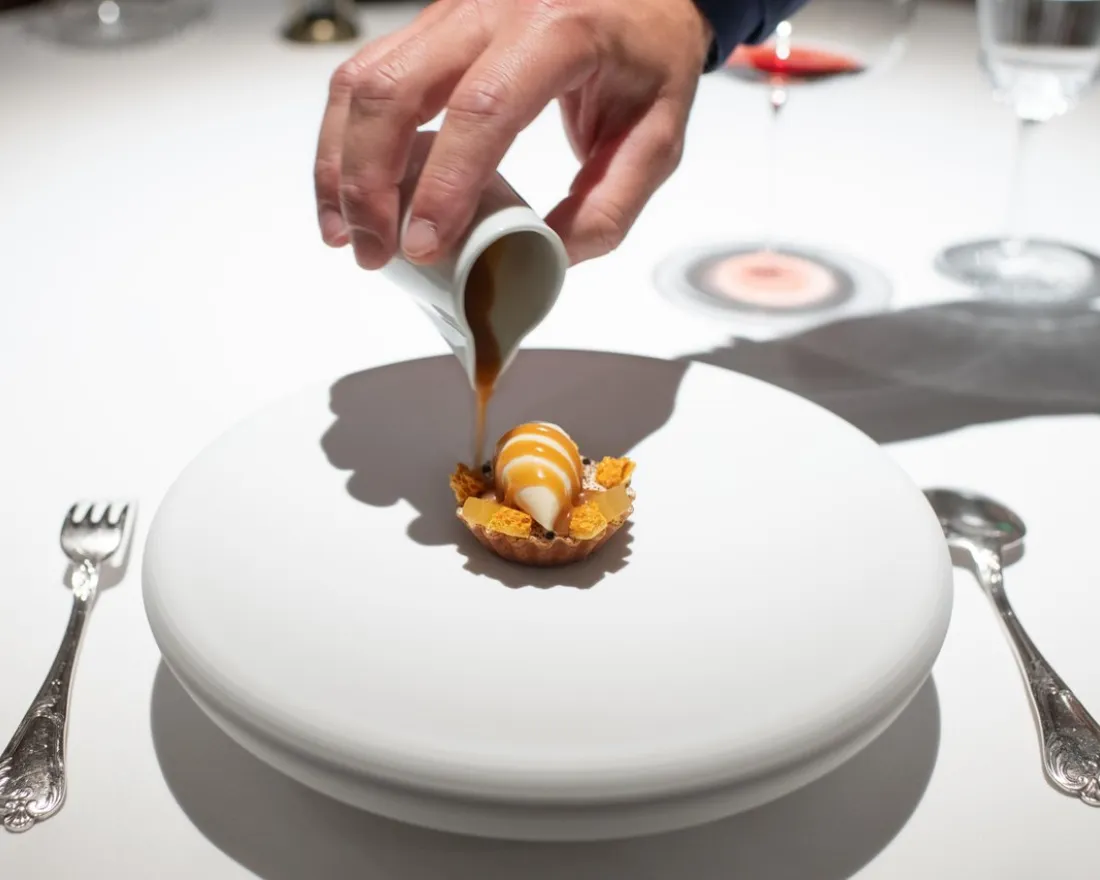 A hand pouring sauce over a dessert on a white plate, with silver utensils nearby.