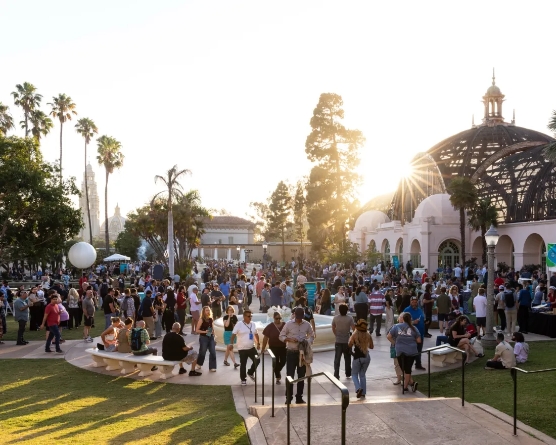 Crowd of people gathered in a Balboa Park during sunset, with buildings and palm trees in the background.