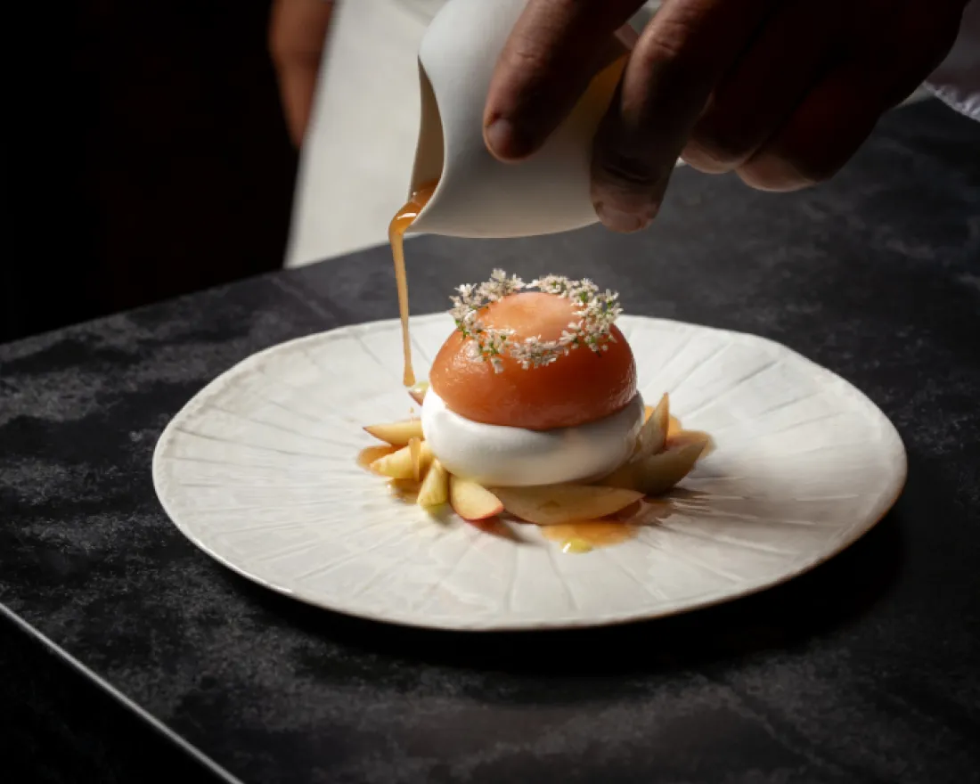 A hand pours sauce over a beautifully presented dessert on a white plate.