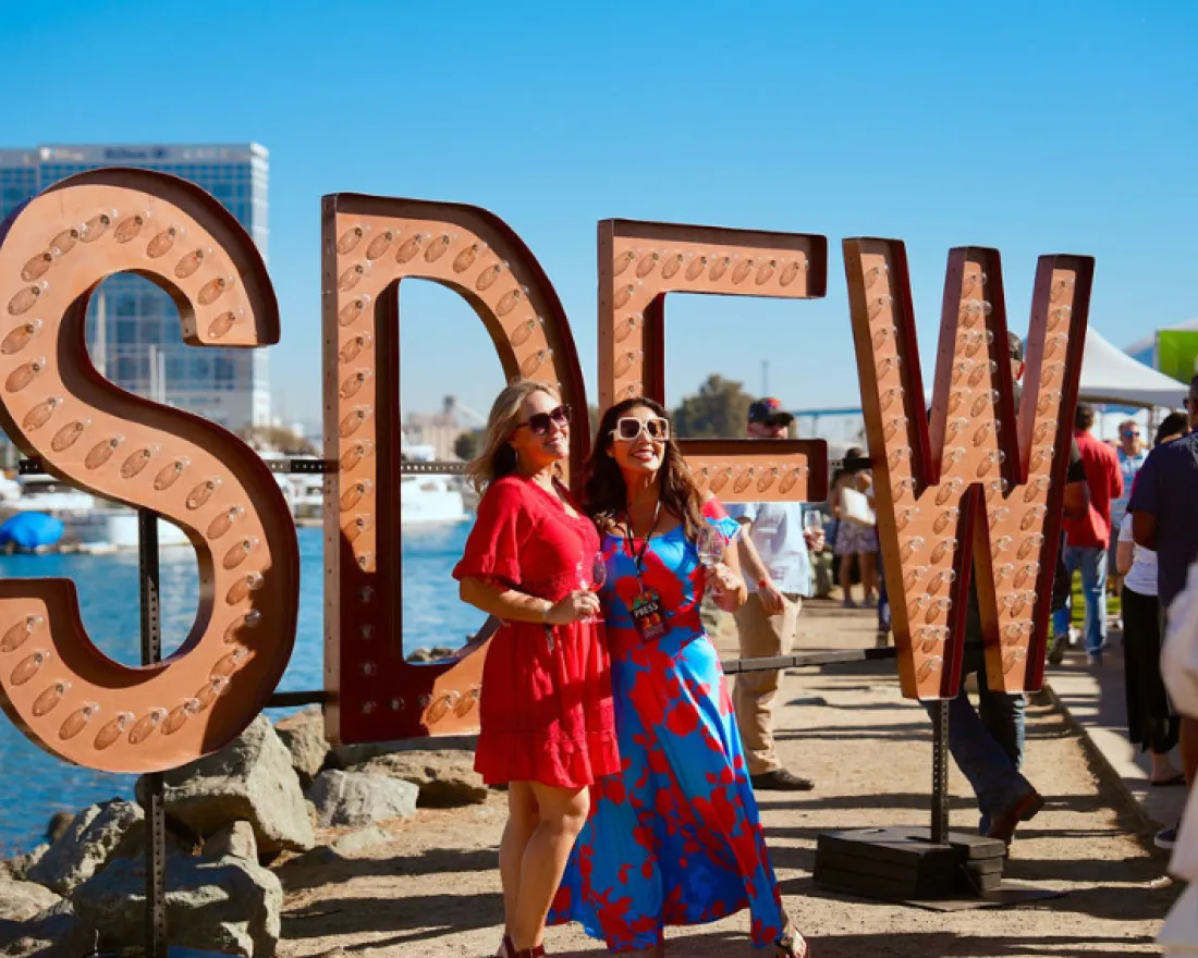 Two women pose in front of large illuminated letters spelling "SDEW" by a waterfront.